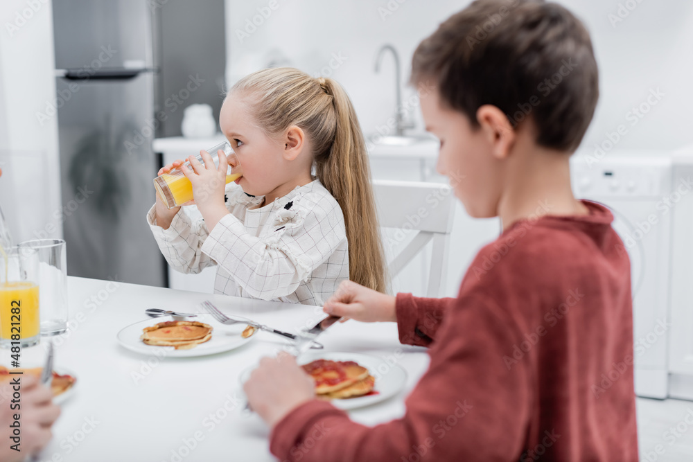 girl drinking orange juice near pancakes and blurred brother.