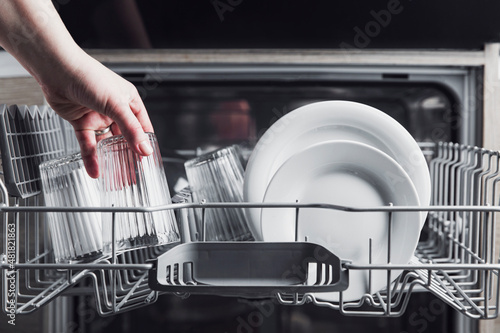 Female hand loading dished, empty out or unloading dishwasher with utensils. Kitchen appliances, lifestyle view. Woman puts a plate in the dishwasher or takes from it. Housewife does her housework 