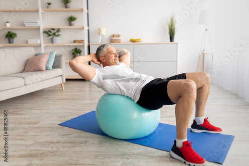 Domestic workout concept. Sporty middle-aged man doing abs exercises at home, leaning on swiss fitness ball photo