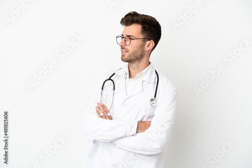 Young doctor caucasian man over isolated on white background wearing a doctor gown and with arms crossed looking side