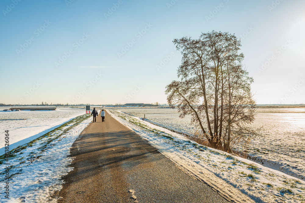 A man and a woman are walking together on a country road in a Dutch polder. It is winter, the sun is low and the fields are covered with snow. A man and a woman are walking together on a country road in a Dutch polder. It is winter, the sun is low and the fields are covered with snow.