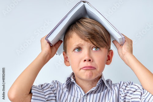 Crying Caucasian School Child Boy Carrying Book on Head on White Studio Background, Copy Space. Sad Upset Kid Depressed By Going Back to School. Motivation, Education, Childhood Concept photo