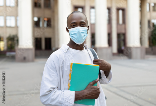 African american male student in face mask standing outdoors with notebooks, posing to camera near university building © Prostock-studio