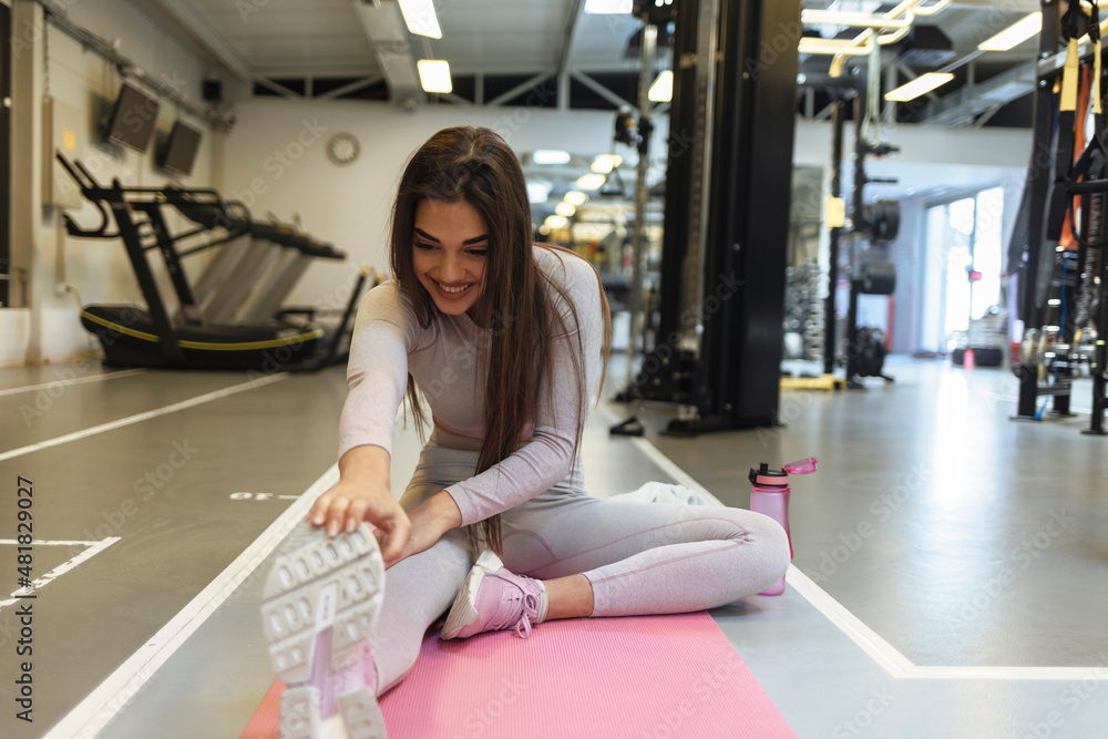 Obraz premium Image of gorgeous young fitness woman sitting in gym near exercise machines while make stretching exercises. Image of strong sports women make sport stretching exercises in gym. Front view.