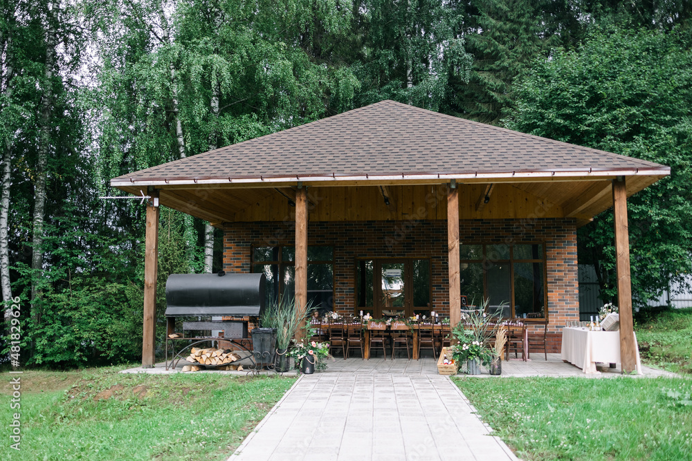 A large wooden gazebo for relaxing. Wedding dinner in nature. Large grill. there is a banquet wooden table decorated with compositions, glasses, candles and plates are placed on the table. Kross back 