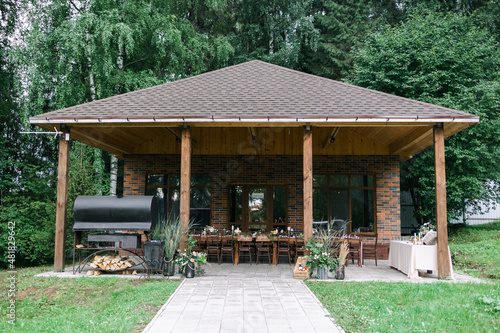 A large wooden gazebo for relaxing. Wedding dinner in nature. Large grill. there is a banquet wooden table decorated with compositions, glasses, candles and plates are placed on the table. Kross back 