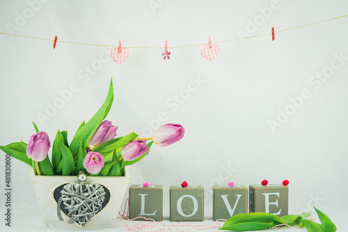 Walentynkowe tło, różowe tulipany w pudełku drewnianym z serduszkiem i napis z brył drewnianych układające się w sowo love, zawieszone na sznurku serduszka.