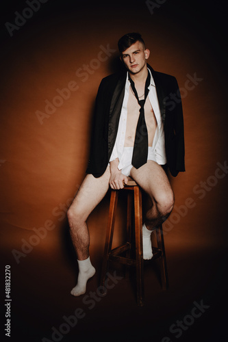 Boudoir portrait. Handsome sexy young brunette male model dressed in black suit, white underwear, shirt and socks sitting on high wooden stool; on brown background. Male beauty concept