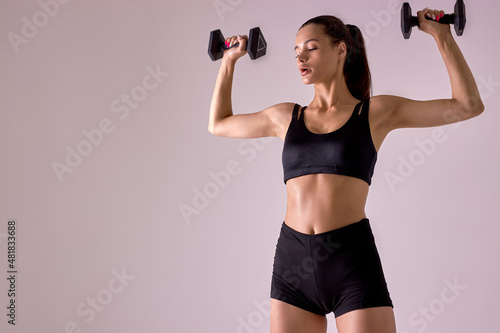 Attractive young brunette woman wearing black sport clothes exercising with dumbbells doing fitness workout, weightlifting, weight loss. portrait of slim lady engaged in sport, raising hands up