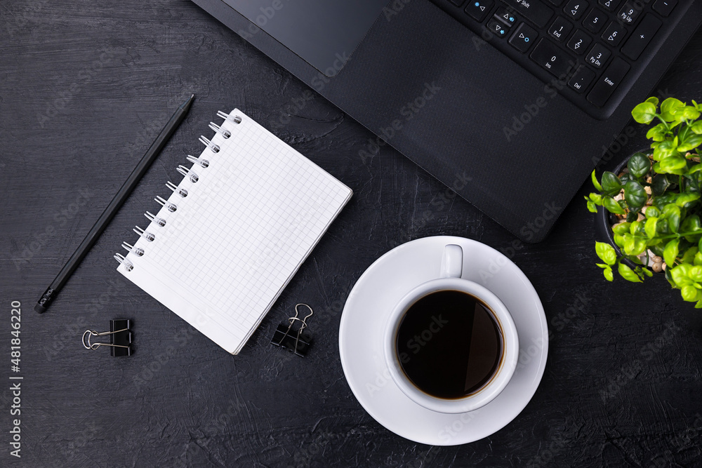 A black laptop, a white checkered notebook and a black pencil on a black table. Black coffee in a white cup and a green potted plant in the background. Top view A black laptop, a white checkered notebook and a black pencil on a black table. Black coffee in a white cup and a green potted plant in the background. Top view