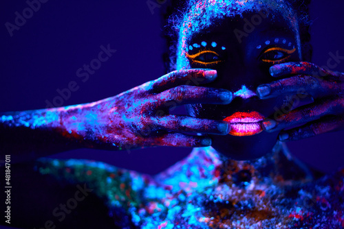 young black woman with fluorescent prints on skin, cosmic paint glowing on neon lights, black background in studio. female with body art closing half of face, touching. beauty, fashion concept