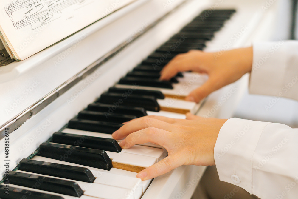 Fototapeta premium Close-up hands of unrecognizable musician female pianist playing gentle music on white classical piano in light classroom. Closeup view of woman music composer practicing piano at home studio.