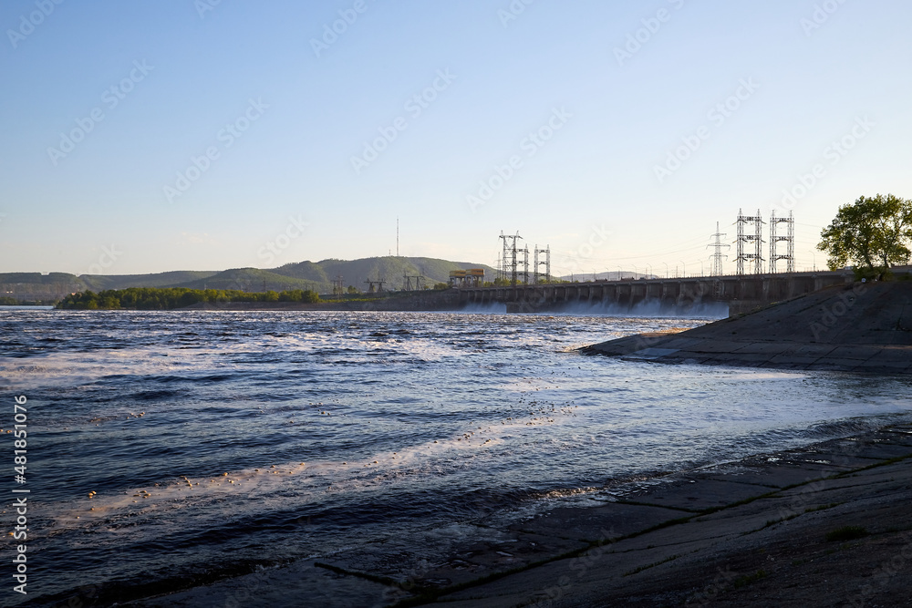 Dam of a reservoir with falling water and a wave on a summer or spring day. Nature landscape with beautiful water. Sea, river, lake, reservoir with blue waves. White foam on the wave