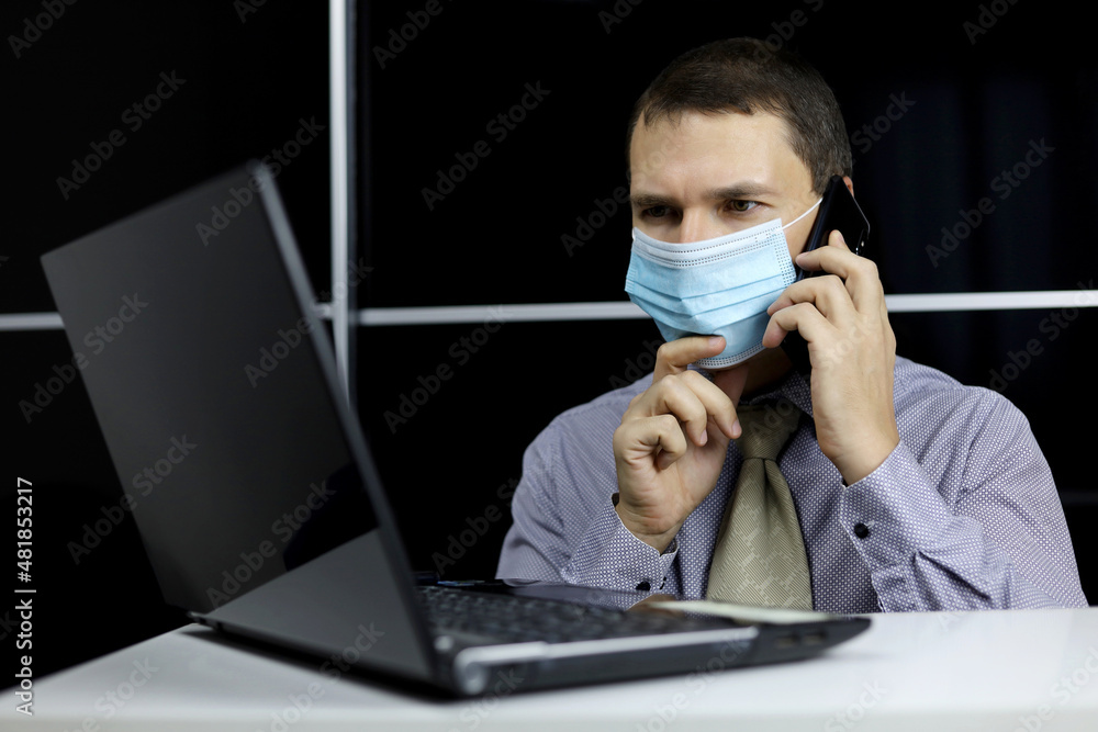 Man in face mask talking on mobile phone sitting at laptop with thoughtful face expression. Discussion of working issues, office call Man in face mask talking on mobile phone sitting at laptop with thoughtful face expression. Discussion of working issues, office call