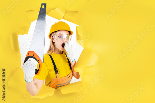 Energy fun handyman blond woman in protective helmet is holding manual saw in torn hole of yellow background. Female in male hard work. Renovation, repair, building concept. Emotional portrait. photo