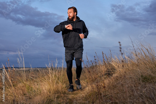 man jogging along field, wearing sportive clothes, practice running outdoors in the morning at sunrise, in nature. young male engaged in sport, motivated, check the time. strong and healthy person