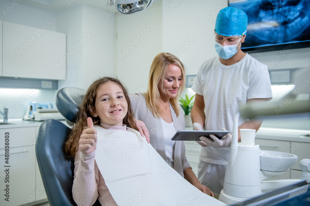 Obraz premium A cute smiling Caucasian little girl looking at the camera showing thumbs up while sitting in a dental chair. In the background, a male dentist and a girl's mother talking and looking at a tablet.