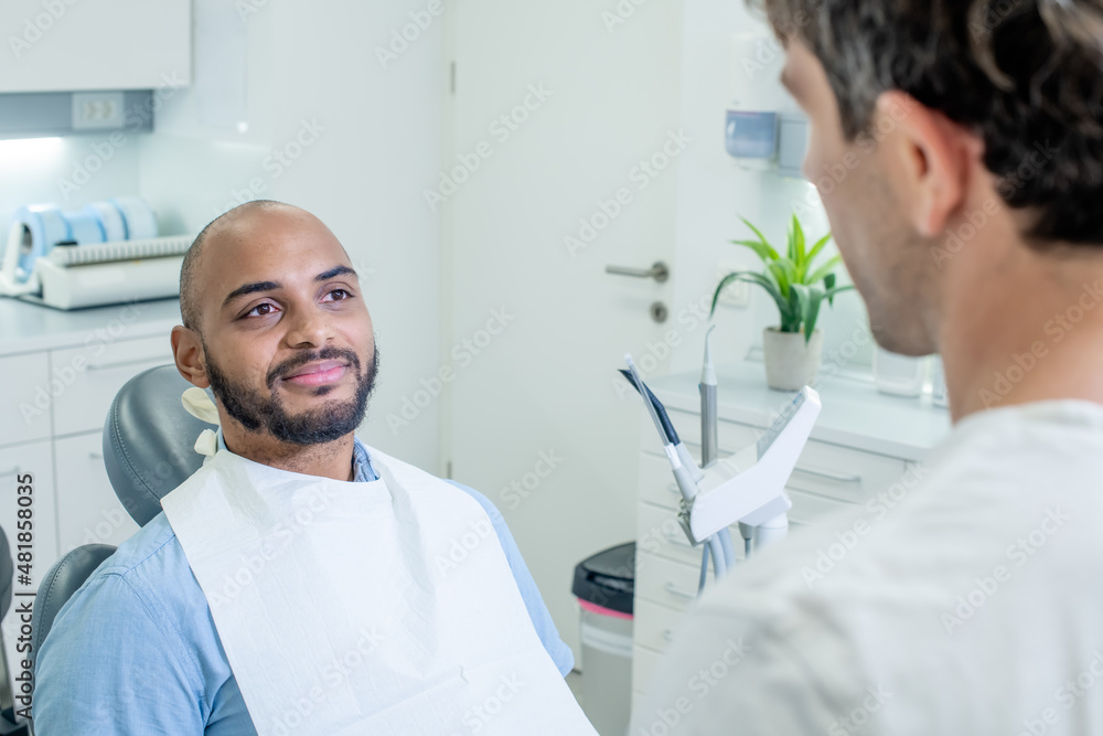 Obraz premium Over the shoulder shot of a male dentist talking reassuringly to his African American male patient, who smiling and looking relaxed while sitting in the dental chair.