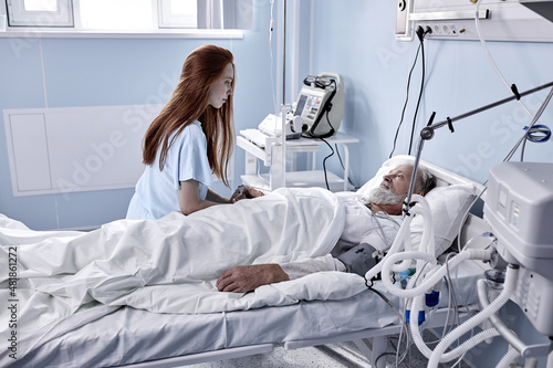 In Hospital Ward Recovering Father is Visited by Adult Daughter. Senior Sick Man Sleeping in Bed, Redhead caucasian Daughter Stands Beside, Worrying. Modern Private Ward. Family Values.