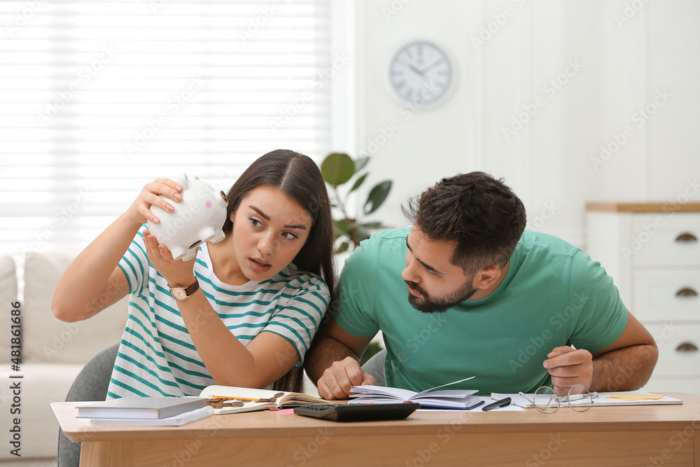 Young couple with piggy bank counting money at wooden table indoors Young couple with piggy bank counting money at wooden table indoors