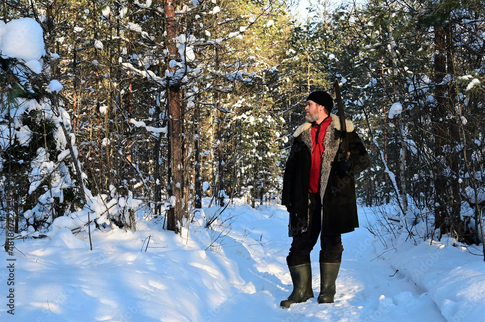 Naklejka premium Portrait of brutal man with beard and in warm unbuttoned sheepskin coat on sunny winter day in snow-covered forest. Man holds hand saw
