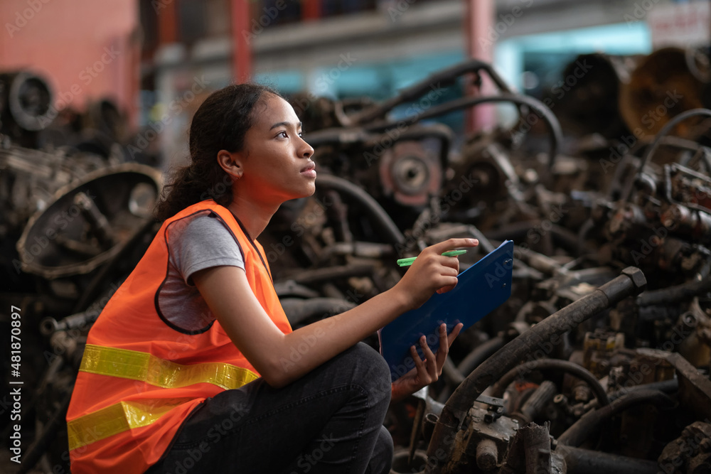 Africa American worker woman standing in the automotive engine part at the factory with copy space. Holding clipboard and checking inventory of old engine in garage. Industrial and stock concept. Africa American worker woman standing in the automotive engine part at the factory with copy space. Holding clipboard and checking inventory of old engine in garage. Industrial and stock concept.