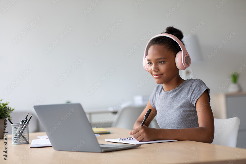 Happy adolescent afro american female student with headphones studying at home watching online lesson Happy adolescent afro american female student with headphones studying at home watching online lesson