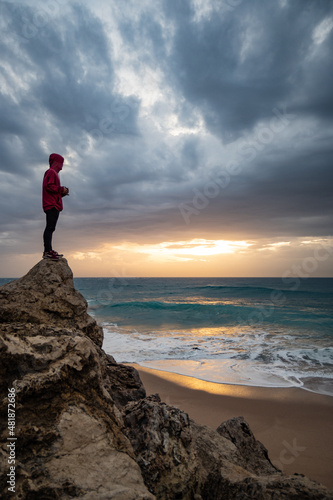 Young man standing on a rock watching waves breaking at the Cape of Trafalgar in a dramatic stormy sunset 