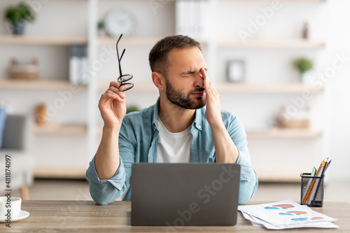 Caucasian male freelancer feeling tired, rubbing irritated eyes, sitting at desk with laptop, exhausted from online work