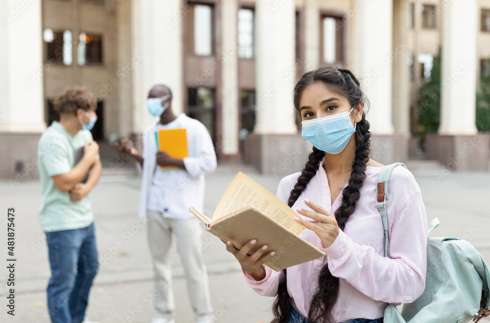 Female student wearing face mask posing with book and looking at camera, standing outdoors near university building Female student wearing face mask posing with book and looking at camera, standing outdoors near university building