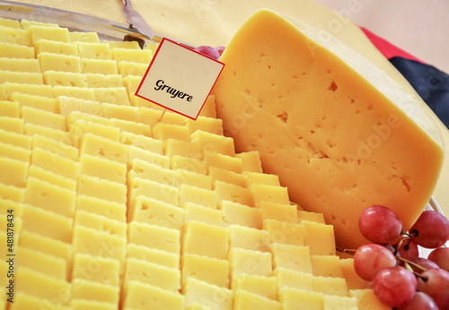 a gruyere cheese, accompanied by cut slices, accompanied by grapes, has a sign that says gruyere.