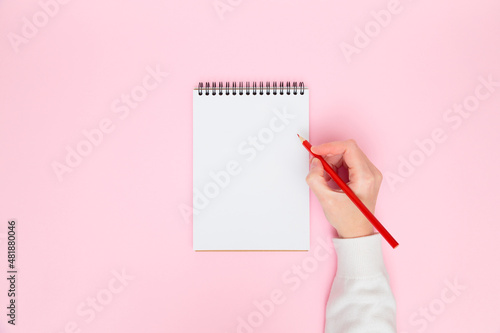 Female hand writing in white blank open spiral notebook with red color pencil isolated on pastel pink background. Flat lay, copy space. Creative work, business, finance or education concept