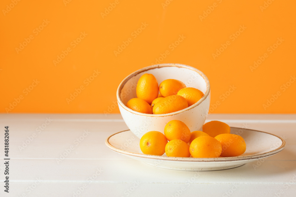 Bowl and plate with tasty kumquat fruits on table against orange wall Bowl and plate with tasty kumquat fruits on table against orange wall