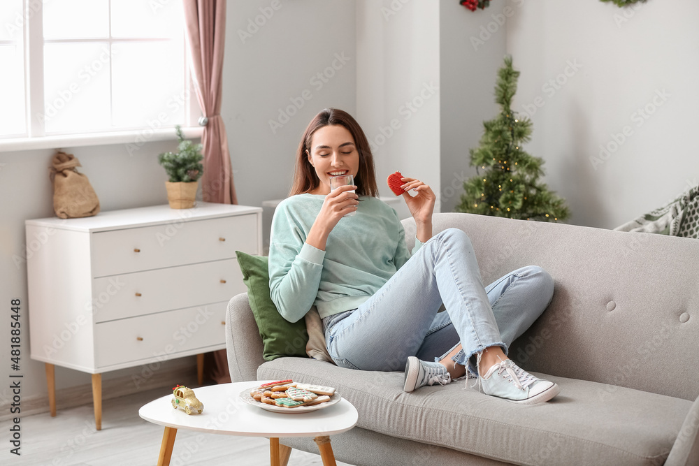 Young woman with gingerbread cookie drinking milk on sofa at home