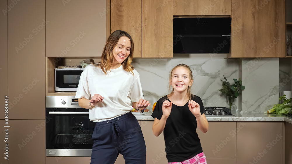 Cheerful long haired mother and teen daughter in black and white t-shirts have fun dancing in kitchen against marble countertop at home. Cheerful long haired mother and teen daughter in black and white t-shirts have fun dancing in kitchen against marble countertop at home.