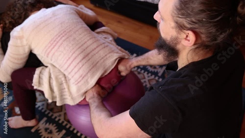 A short clip seen closeup and high angle, as a male therapist applies pressure on the sacroiliac joints of a female client sitting on an exercise ball. photo