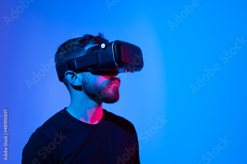 Virtual reality world. Bearded guy in vr headset, touching something while playing video game in metaverse with his friends, standing in neon light.