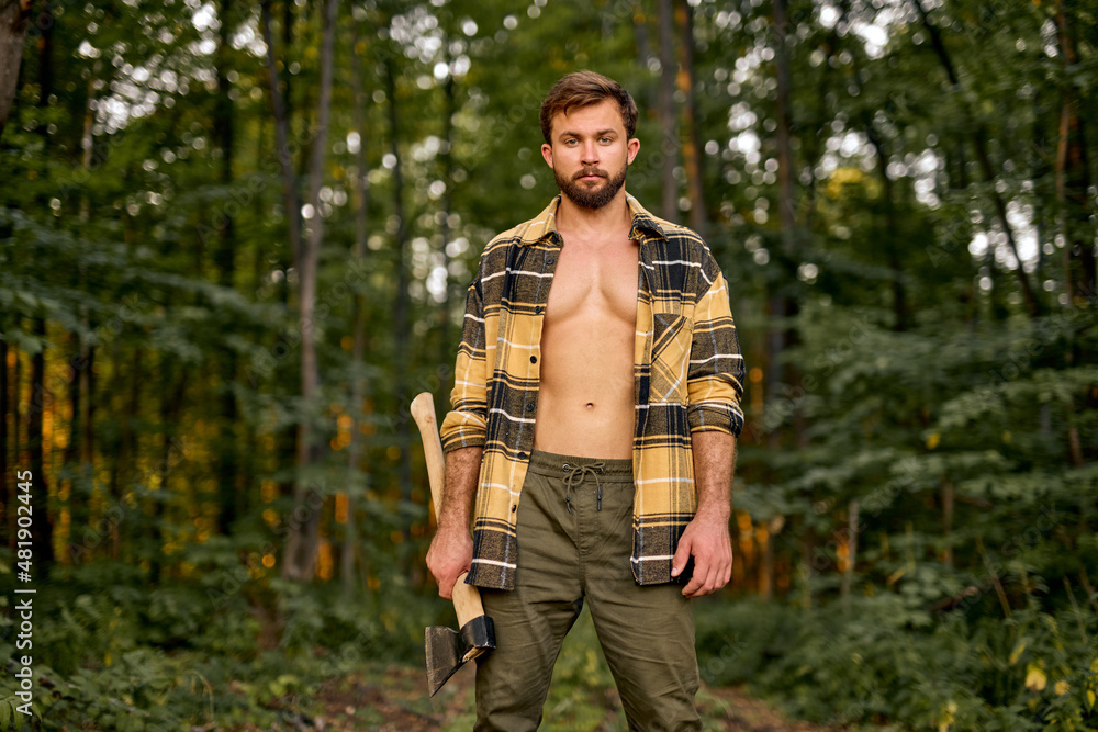 Brutal handsome european man in open plaid shirt carry large splitting axe in summer forest natural landscape, lumberman with perfect muscular body posing at camera, shirtless sexy lumberman