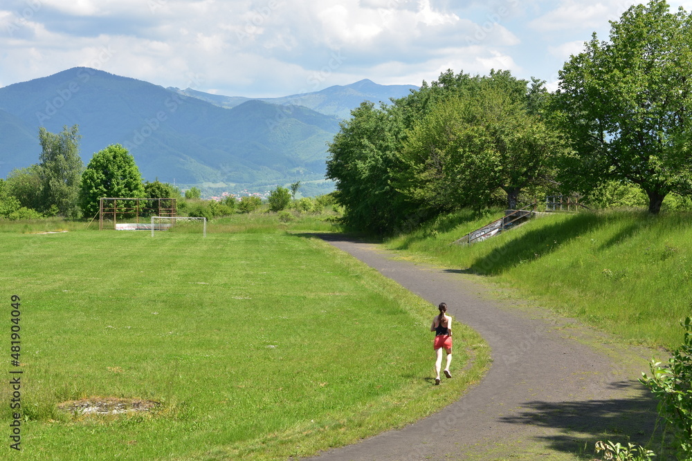 Young long-distance girl runner in red shorts and black sweat shirt running in green grass grown sport athletic stadium. In the background are visible bushes and mountains. Young long-distance girl runner in red shorts and black sweat shirt running in green grass grown sport athletic stadium. In the background are visible bushes and mountains.