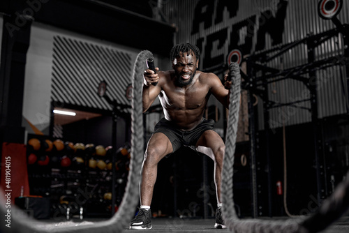 afro american fit athlete doing battle ropes exercise at crossfit gym. African Male wear black shorts training with rope. sport motivation, cross fit, fitness concept. cardio training