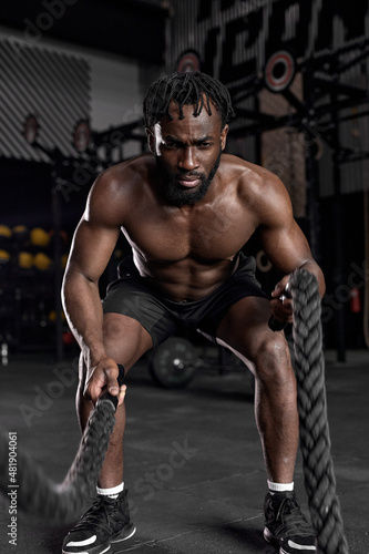 Young black man doing strength training using battle ropes at modern dark gym. Athlete moving the ropes in wave motion as part of fat burning workout, active intense cross fit training, exercising