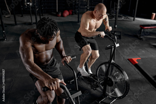 Men exercise bike gym cycling training fitness. Two Fitness male using air bike cardio workout. Two Athlete guys with naked torso biking indoor at gym, exercising legs. Cross functional training.