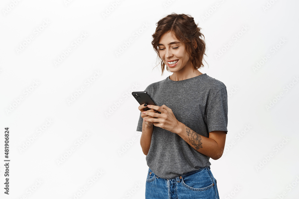 Smiling authentic girl using smartphone app, chatting or ordering, holding mobile phone, standing against white background Smiling authentic girl using smartphone app, chatting or ordering, holding mobile phone, standing against white background