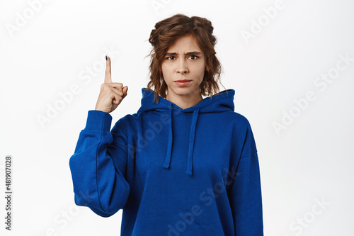 Angry, upset young woman, student pointing finger up and looking disappointed, complaining, sulking at smth unfair, standing over white background