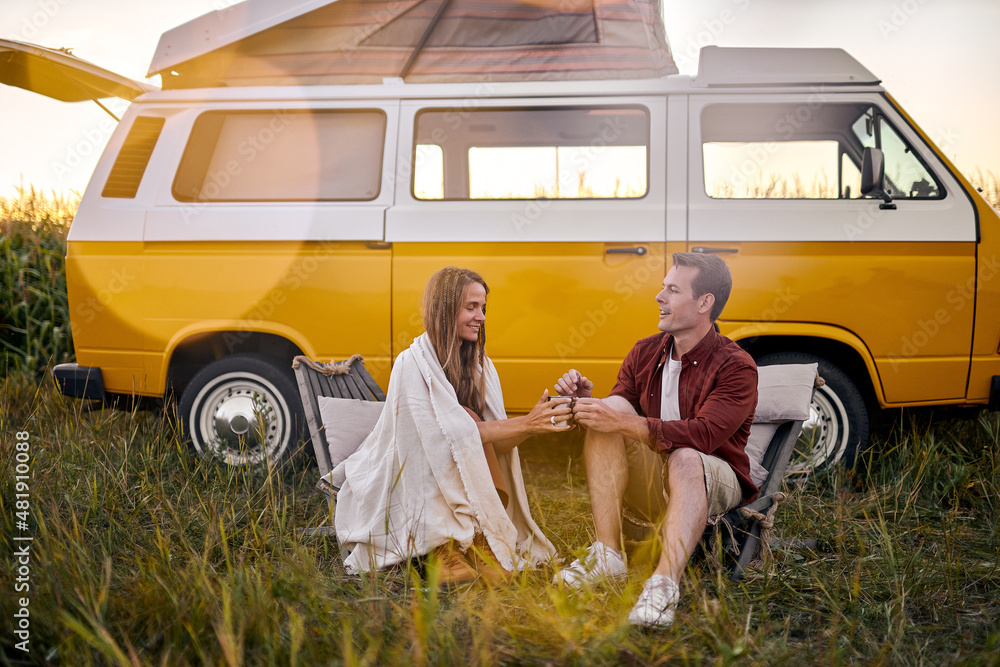 Foto Stock Beautiful happy Hippie couple sitting next to yellow retro style  van vehicle camper trailer, caucasian man and woman drinking cup of tea  from thermos. Female camping, traveling, hitchhiking. | Adobe