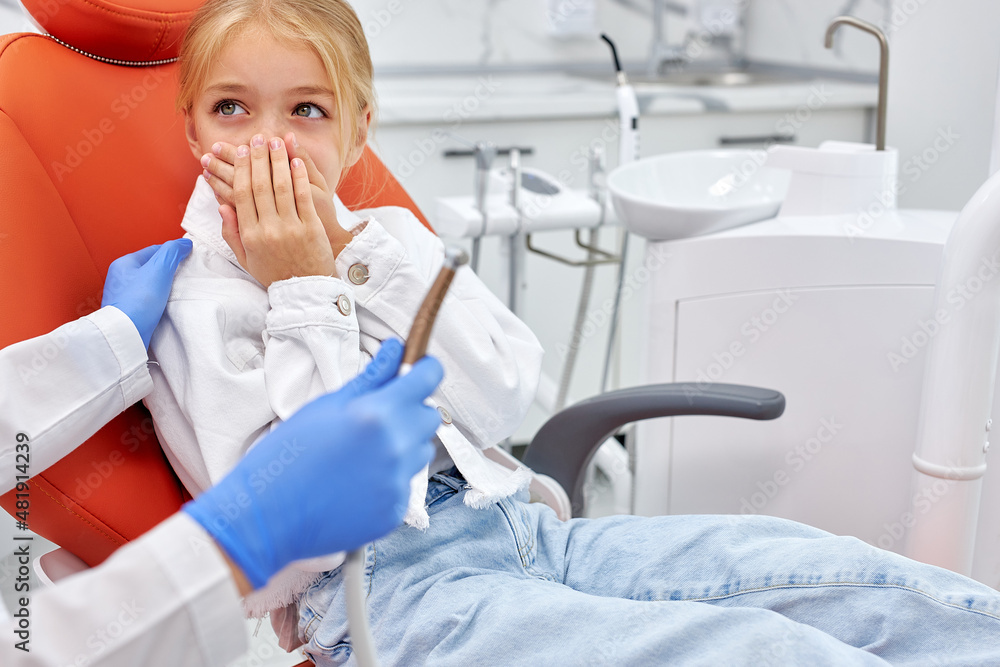 Fototapeta premium Portrait of little cute child closing mouth by fear in dental clinic, frightened by dental drill, afraid of treatment, cropped unrecognizable female doctor in blue gloves holding dental tool in hands