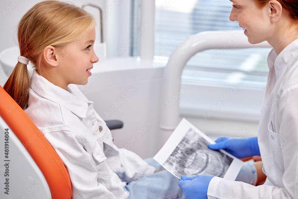 Fototapeta premium Side View On Kid Girl Listening To Female Dentist, Having Talk About Teeth Treatment. Stomatologist Showing X-ray Picture To Child Girl Patient. Medicine, healthcare, treatment concept