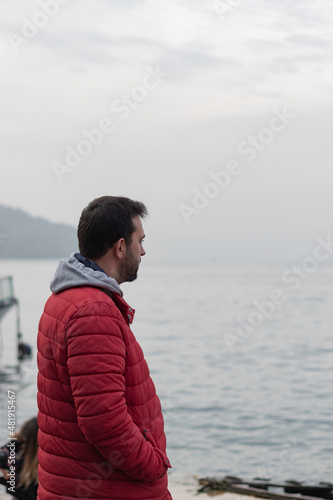 Lonely man standing alone by sea with winter clothes. Hands in their pockets. He watches the waves and the city thoughtfully. Loneliness and dramatic concept photo.