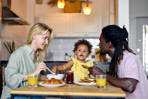 Cute family having breakfast together, enjoy meal at home in kitchen, in the morning. Attractive blonde mother, black dad and black daughter sit behind table eating food. everyday lifestyle concept