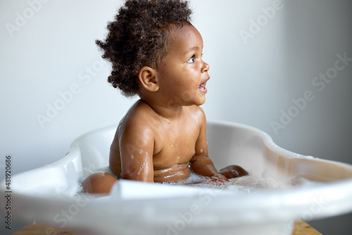 Tableau sur toile Small cute black child sitting at bath, washing, playing with soap and foam, indoors, copy space, white background, happy childhood, banner
